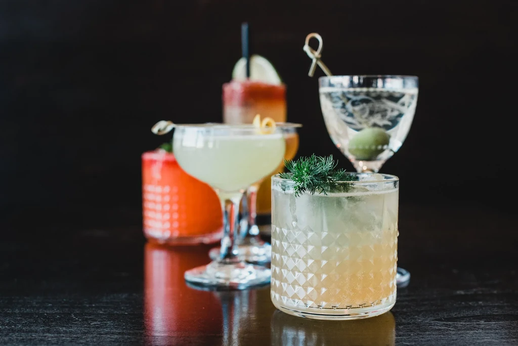 Five colorful garnished cocktails on a dark reflective surface.