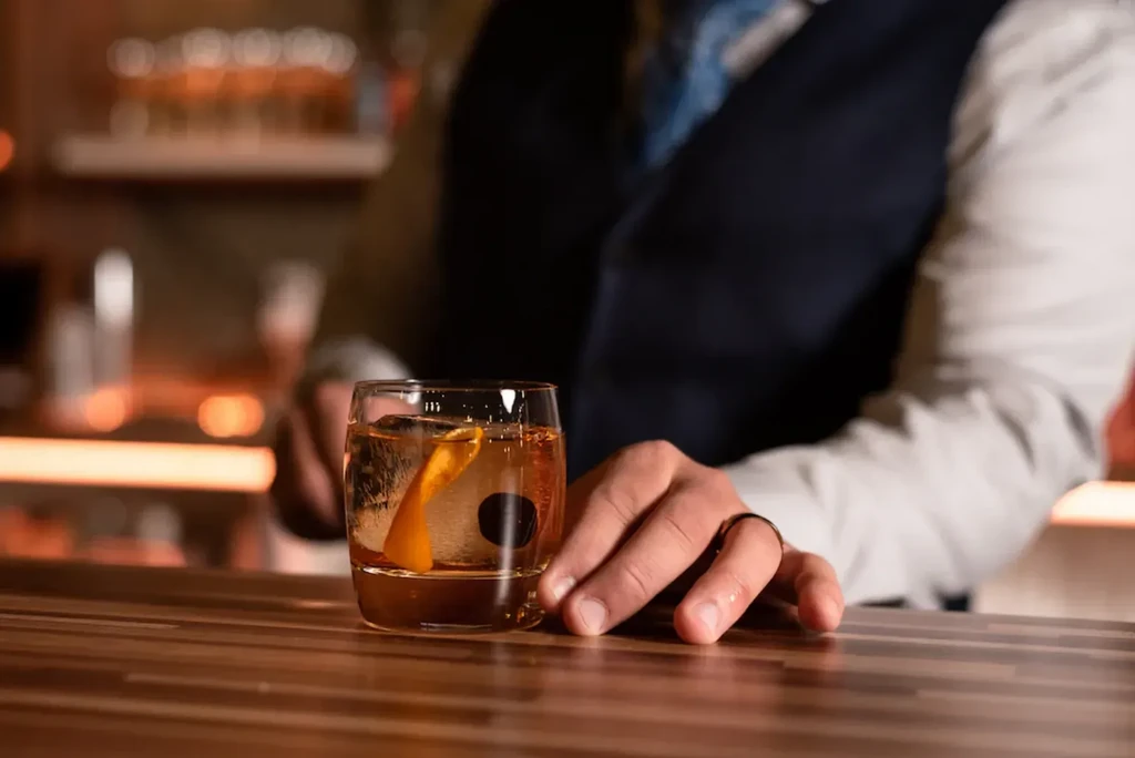 Amber cocktail with ice and orange peel on bar, hand resting nearby.