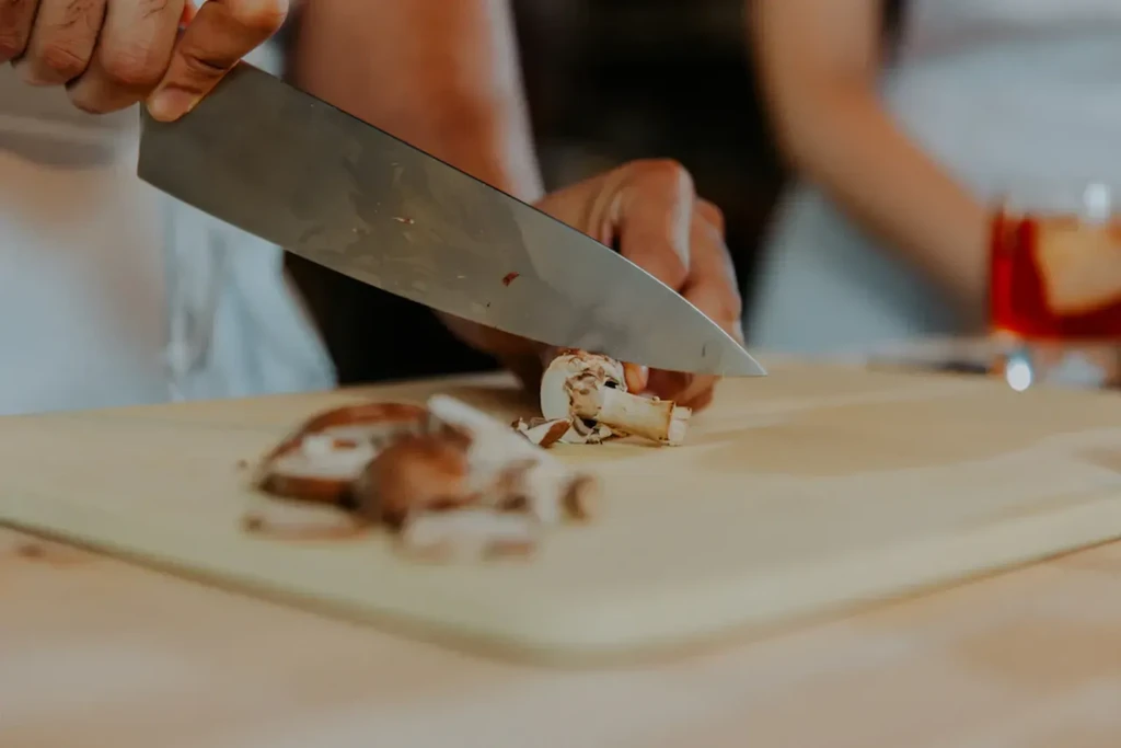 Hands holding large chef's knife, slicing mushrooms on cutting board.