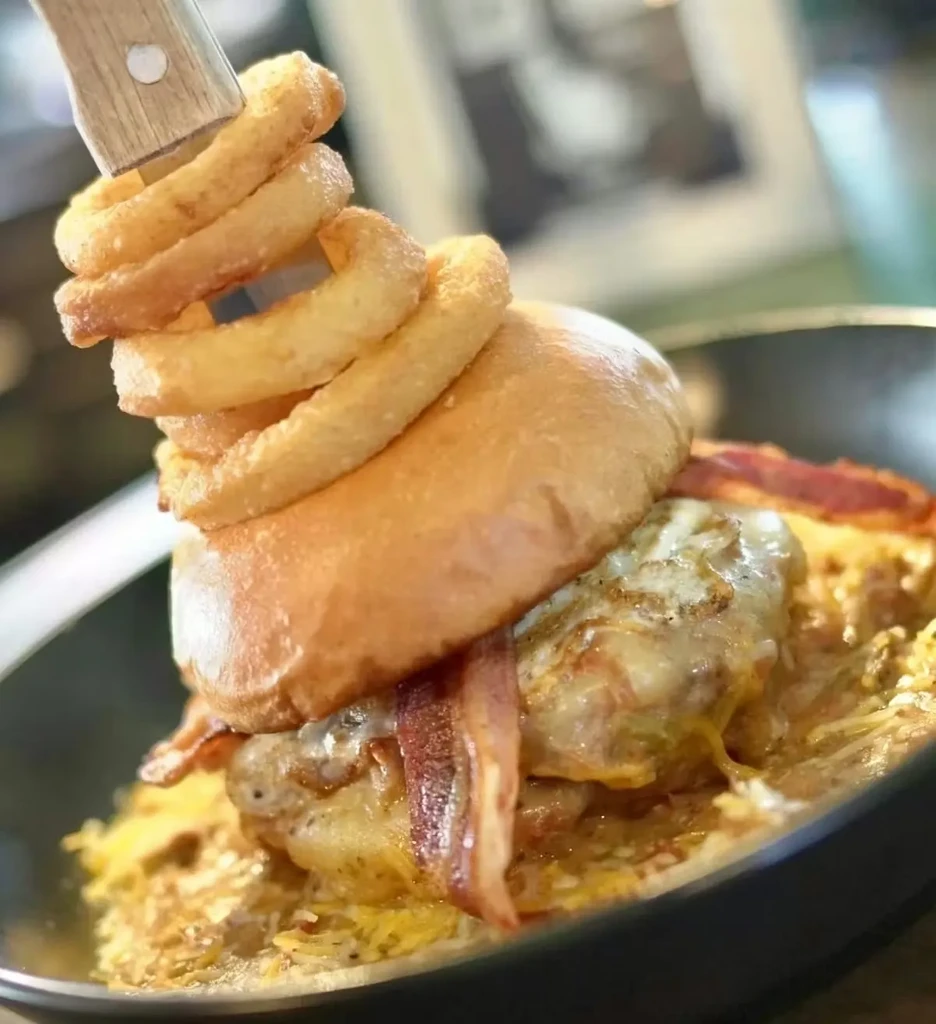 Burger with onion rings, bacon, cheese, skewered by knife in bowl.