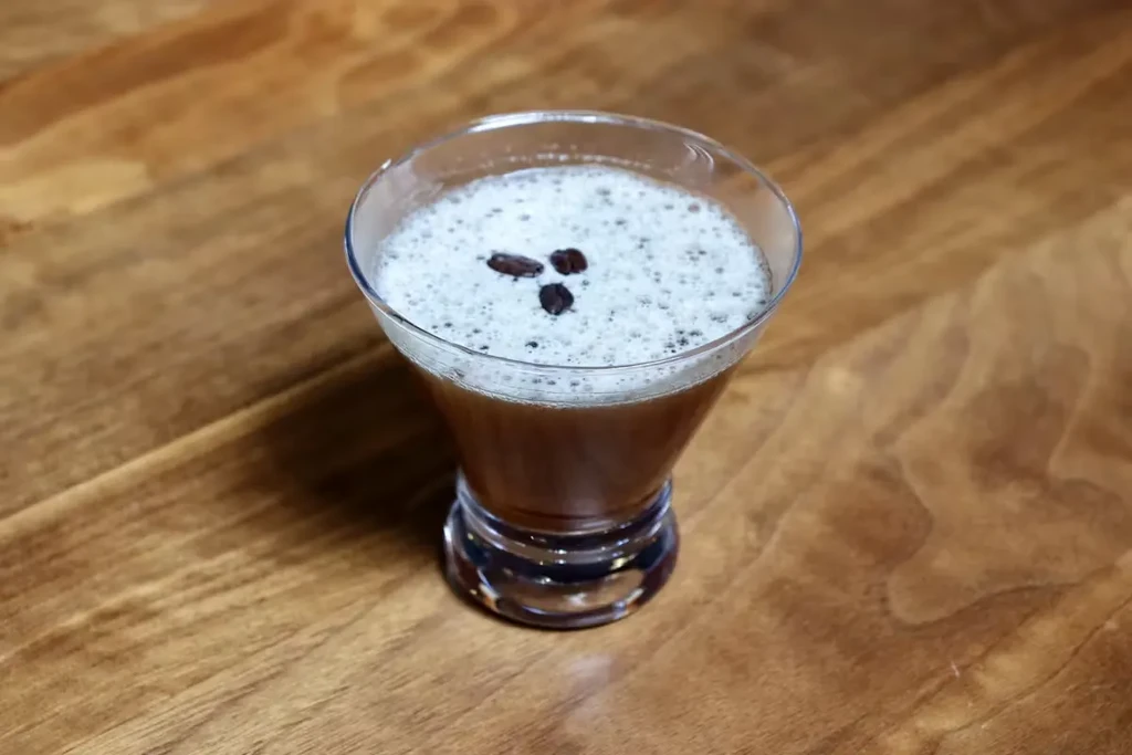 Dark brown cocktail with white foam and three coffee beans on wood.