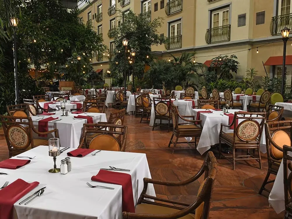 Empty indoor atrium restaurant with lush plants, set tables, and string lights.