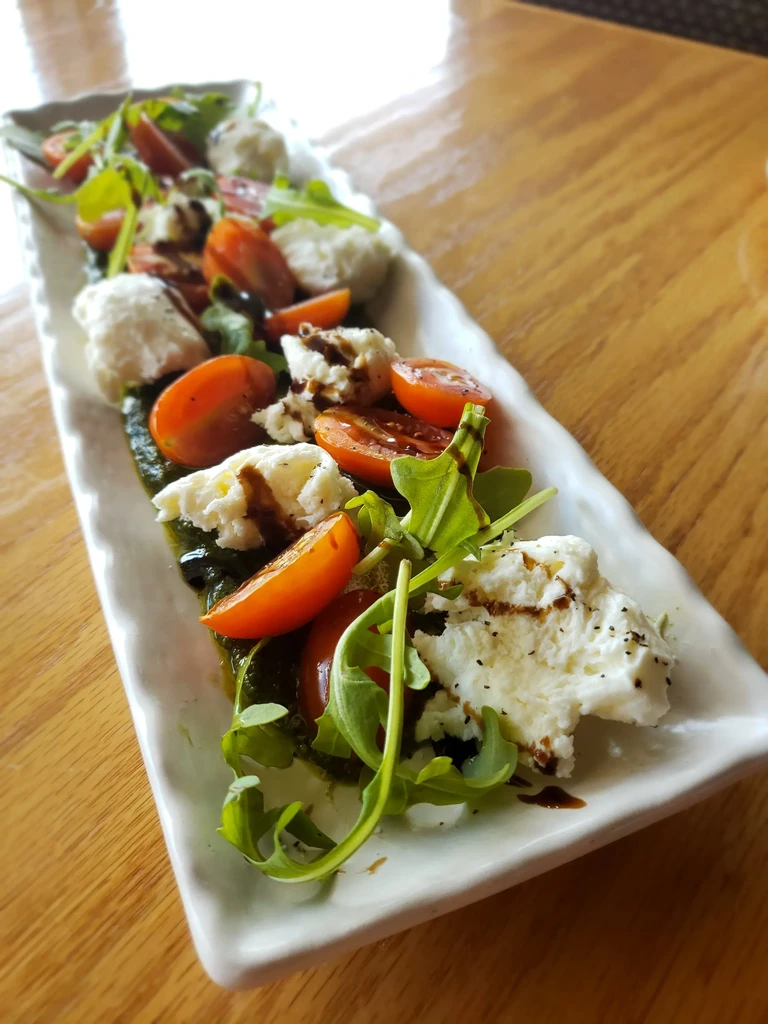 Close-up of deconstructed caprese with mozzarella, cherry tomatoes, arugula, pesto, balsamic.