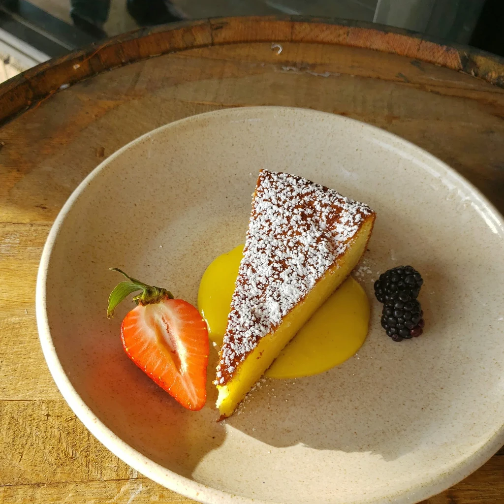 Yellow cake slice with powdered sugar, berries, and sauce on wood.
