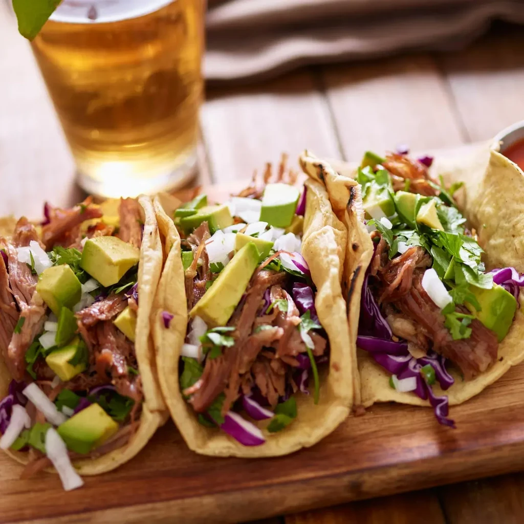 Three street tacos with meat, avocado, onions, cilantro on a wood board.