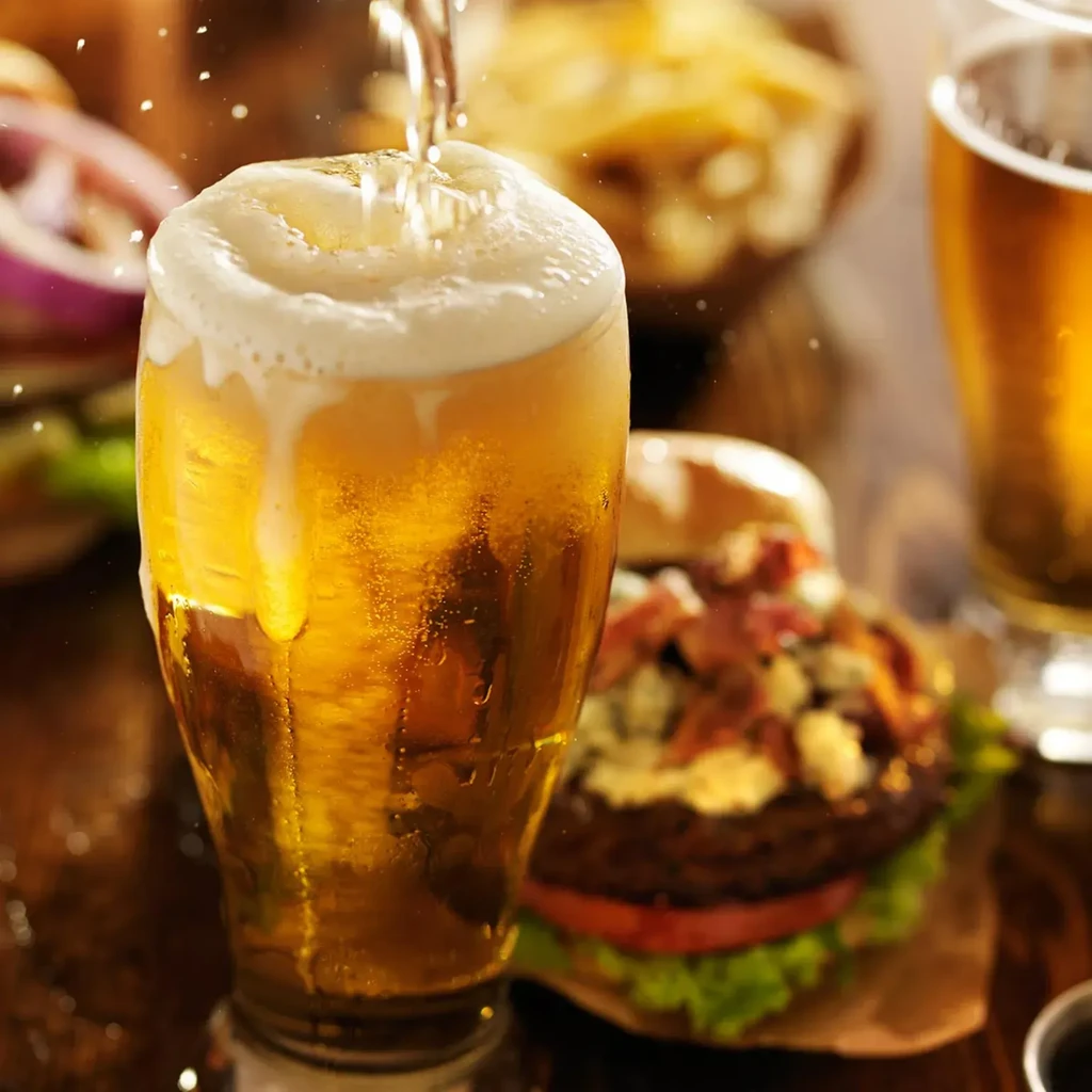 Golden beer pouring into a foamy, cold glass; burger behind.