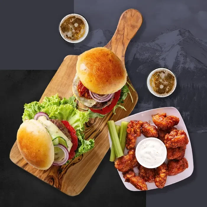 Top-down view: Two burgers, wings, celery, dip, and two beers.