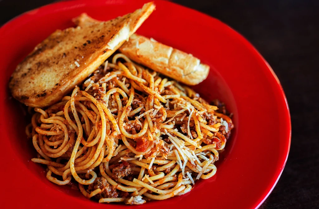 Red plate of spaghetti bolognese, cheese, and two garlic bread slices.