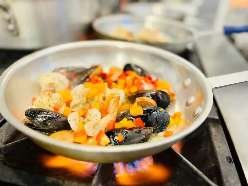 Close-up of stainless steel pan cooking mussels, shrimp, peppers on gas stove.