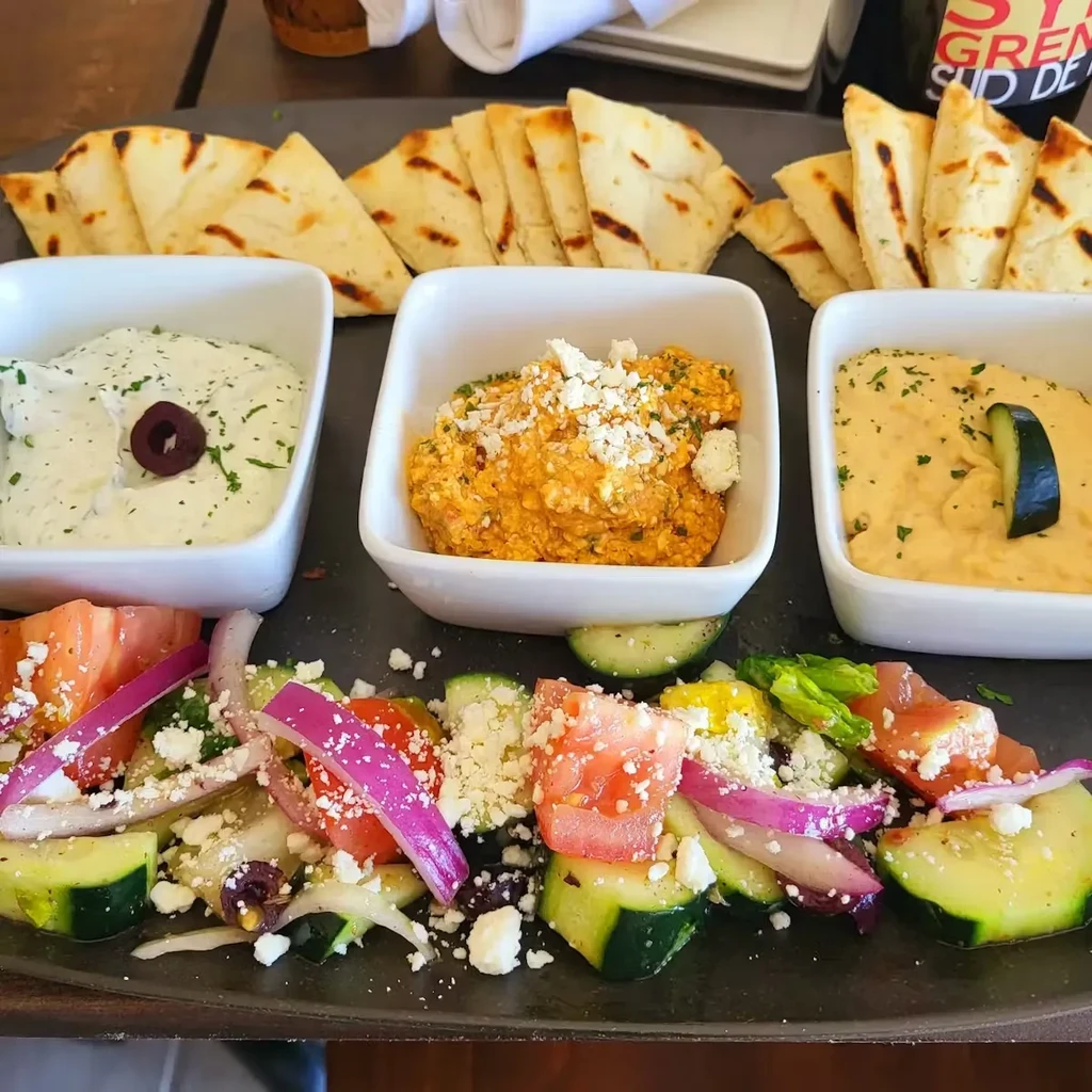 Platter of Greek dips, pita bread, and salad on a dark tray.