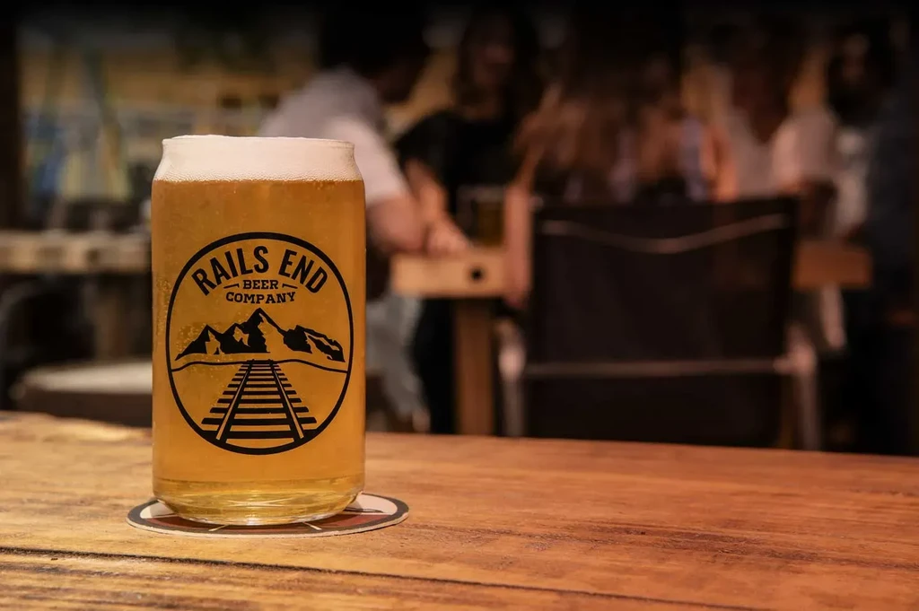 Glass of golden beer with Rails End Beer Company logo, blurred background.