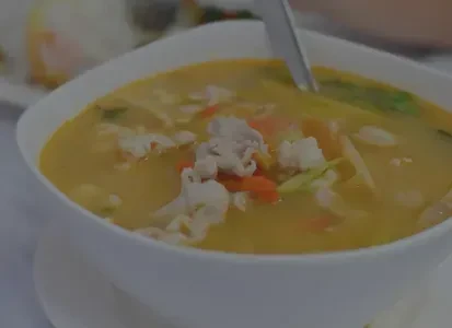 White bowl of yellowish soup with meat, carrots, and leafy greens.