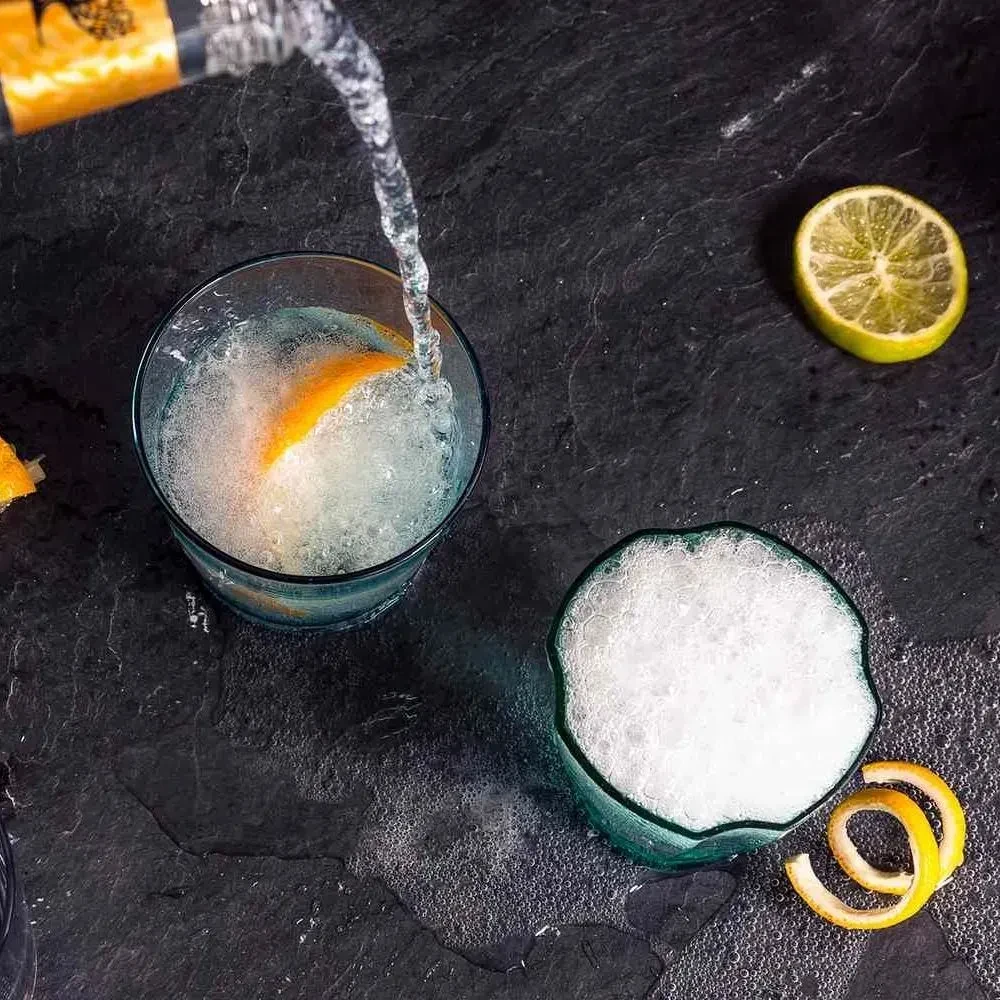 Two drinks with citrus garnishes, one pouring on a dark surface.