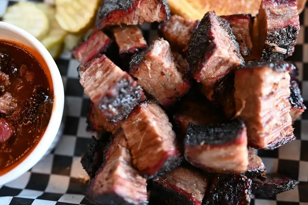 Close-up of BBQ burnt ends, dark sauce, and fries on checkered paper.