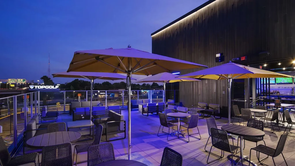 Topgolf outdoor patio with tables, umbrellas, purple lighting at dusk.