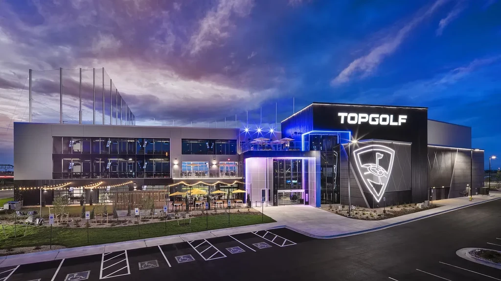 Modern Topgolf complex with neon lights, patio, and golf netting at dusk.
