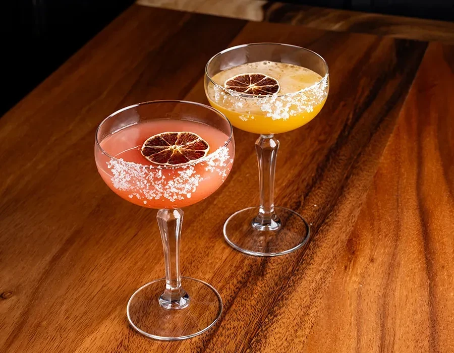 Reddish-pink and golden-yellow cocktails with salt rims on a wooden surface.