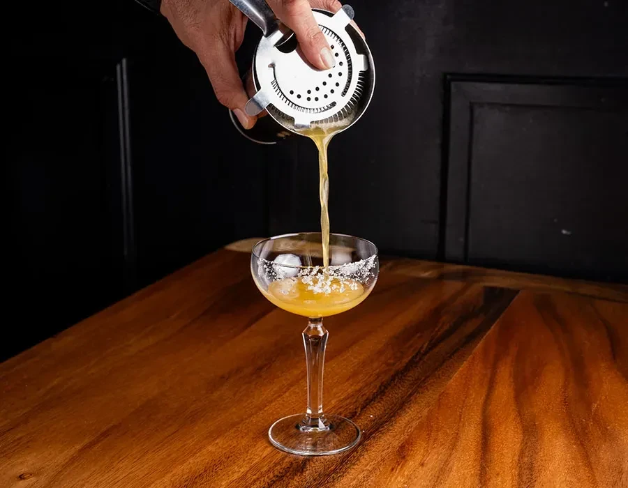 Bartender pours amber cocktail from strainer into sugared coupe glass.