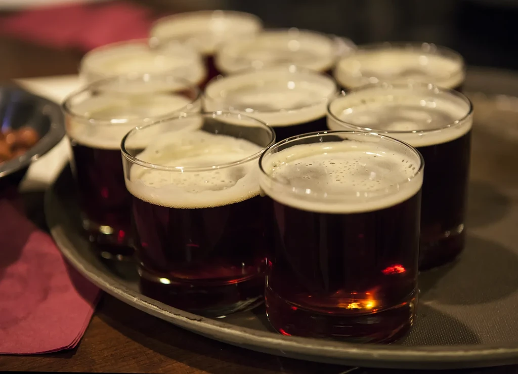Multiple glasses of dark beer with foam and snacks on a tray.
