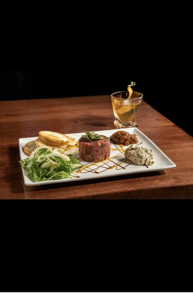 Professionally plated steak tartare with salad, crostini, sauces, and a cocktail.