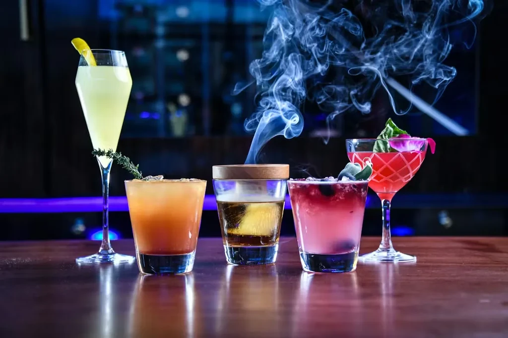 Five vibrant cocktails on a bar, one with dramatic white smoke.
