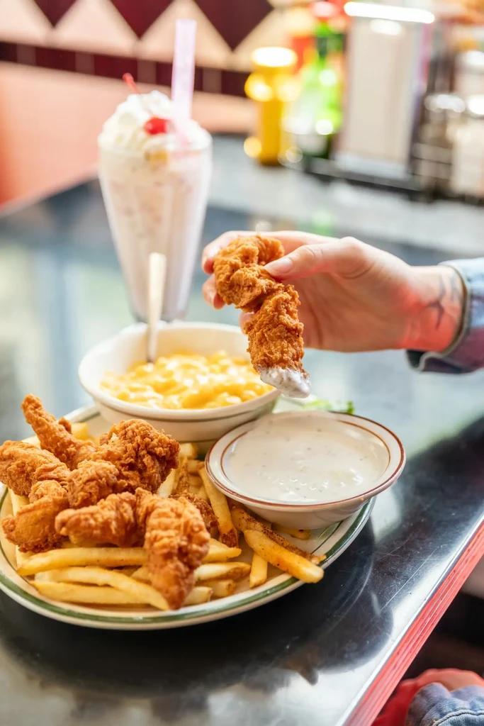 Hand dips fried chicken tender into sauce, with fries and mac.