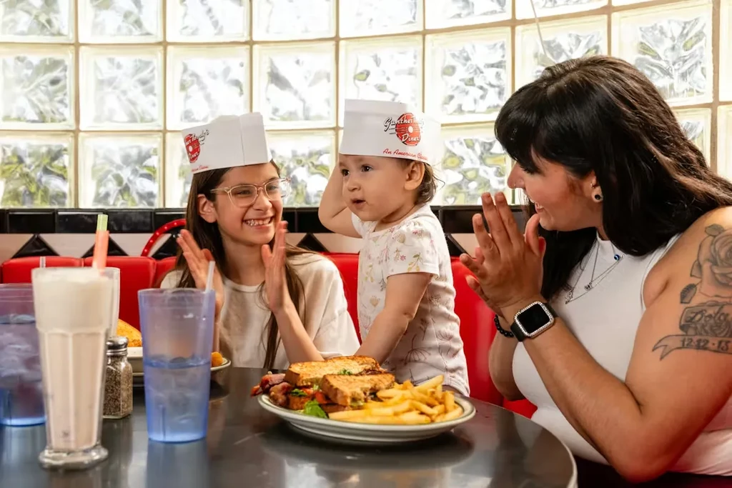 Woman and two children in diner wearing chef hats, clapping.