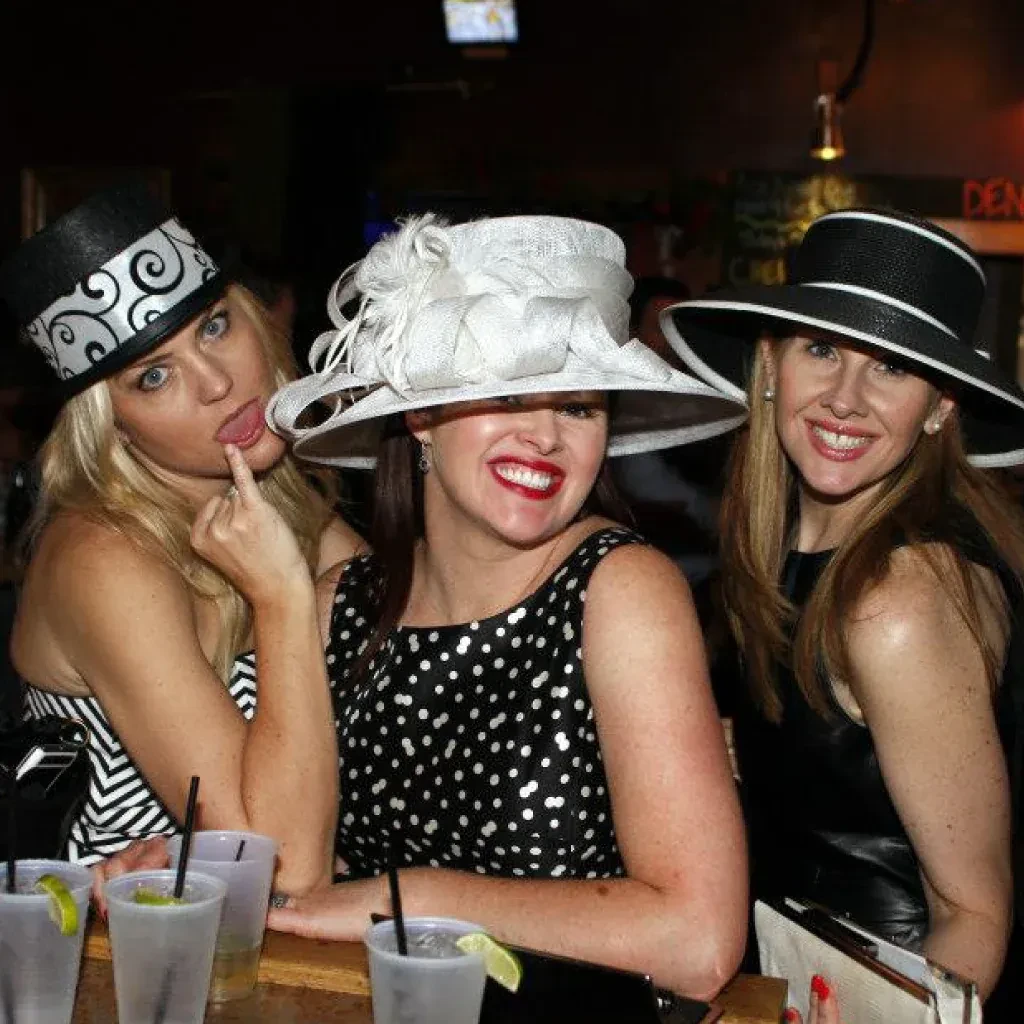 Three women in elaborate hats at a bar, one giving middle finger.
