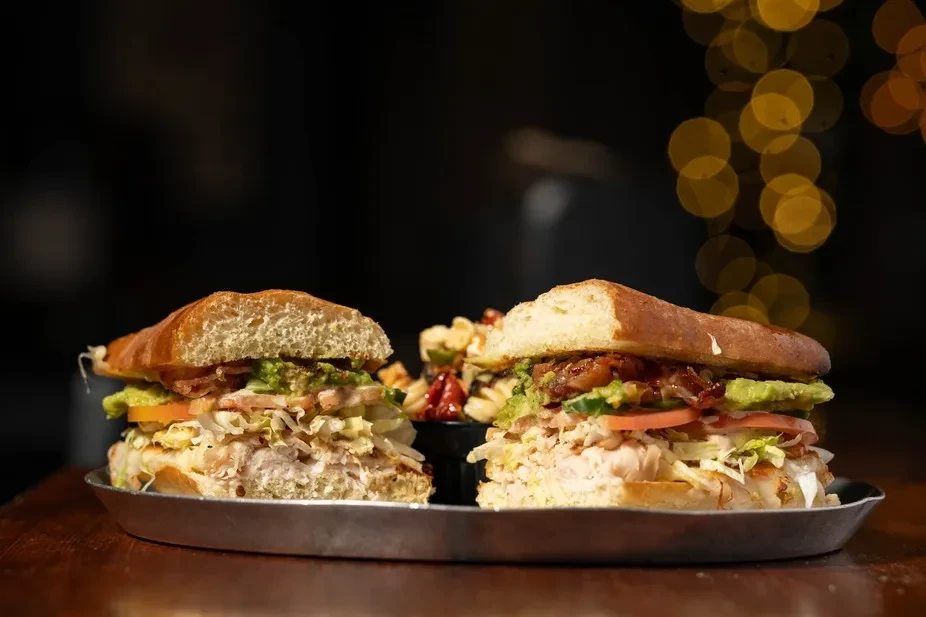 Two large sandwiches on a tray, filled with meat, avocado, bacon, vegetables.