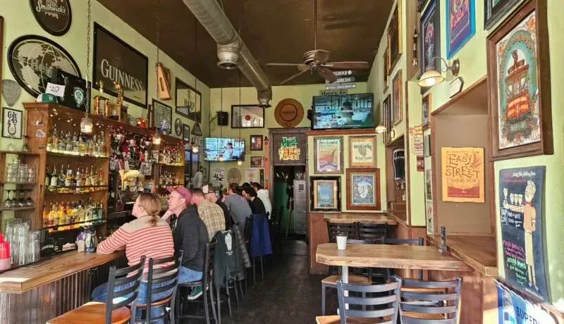 Traditional pub with a bar, patrons, liquor bottles, and empty tables.