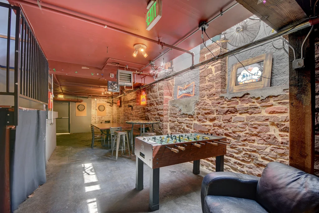 Industrial-rustic bar interior featuring a foosball table, dartboards, and neon signs.