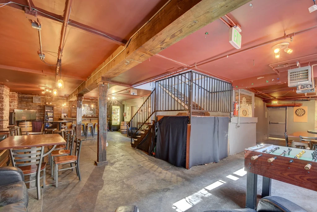 Rustic industrial restaurant interior with exposed beams, bar, staircase, and games.
