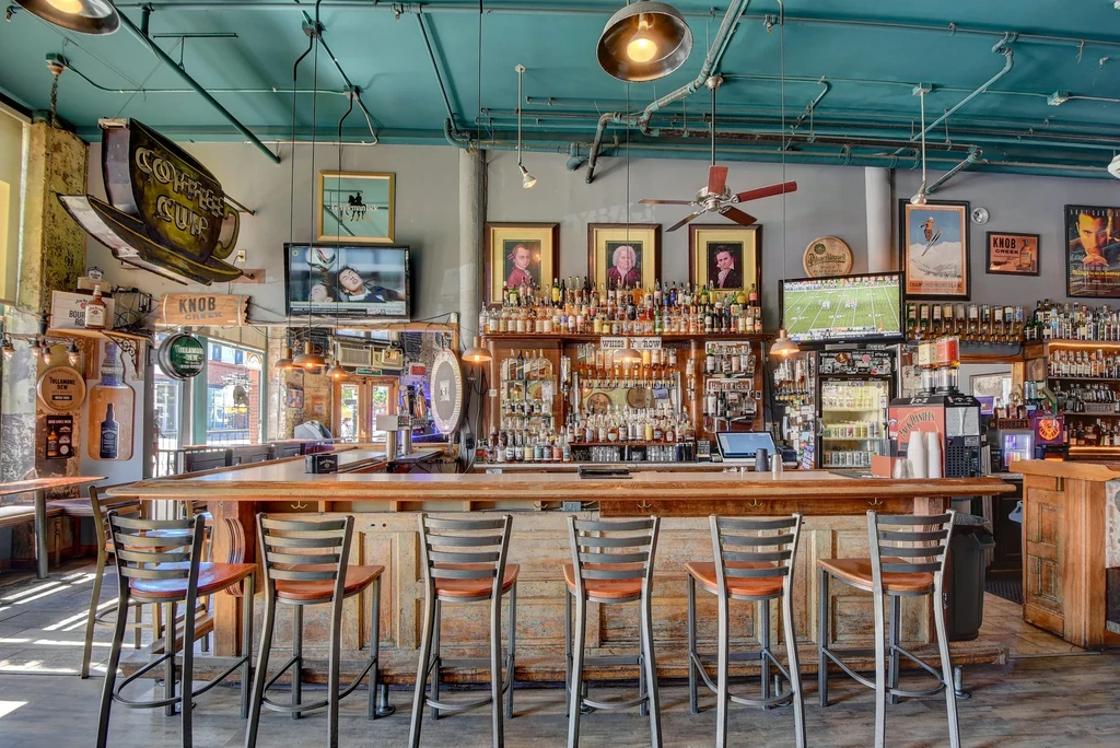 Rustic bar interior with wooden counter, spirits, portraits, and industrial ceiling.