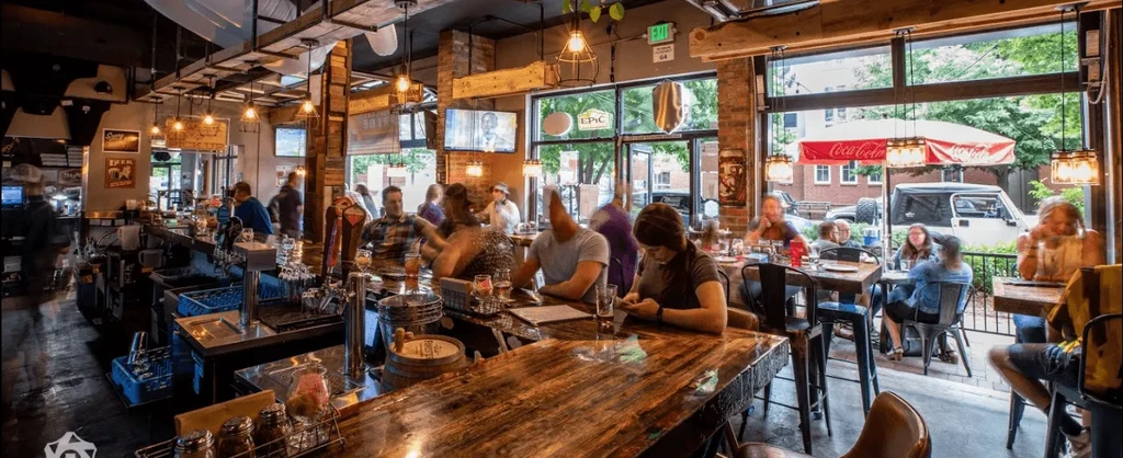 Panoramic, rustic-industrial restaurant interior with a busy bar and diners.