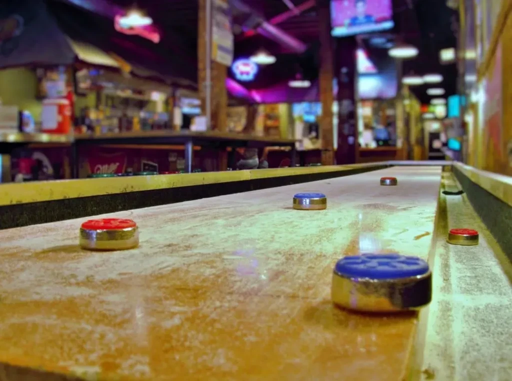 Textured shuffleboard table with red and blue pucks in a blurred bar.