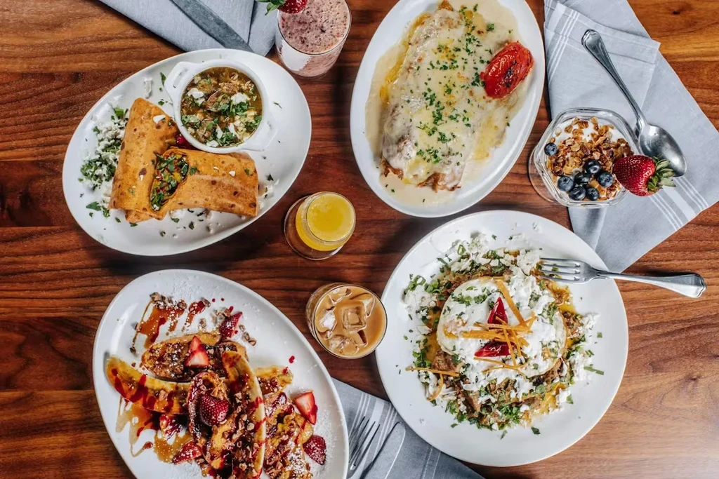 Overhead: wooden table with diverse breakfast and brunch dishes and drinks.