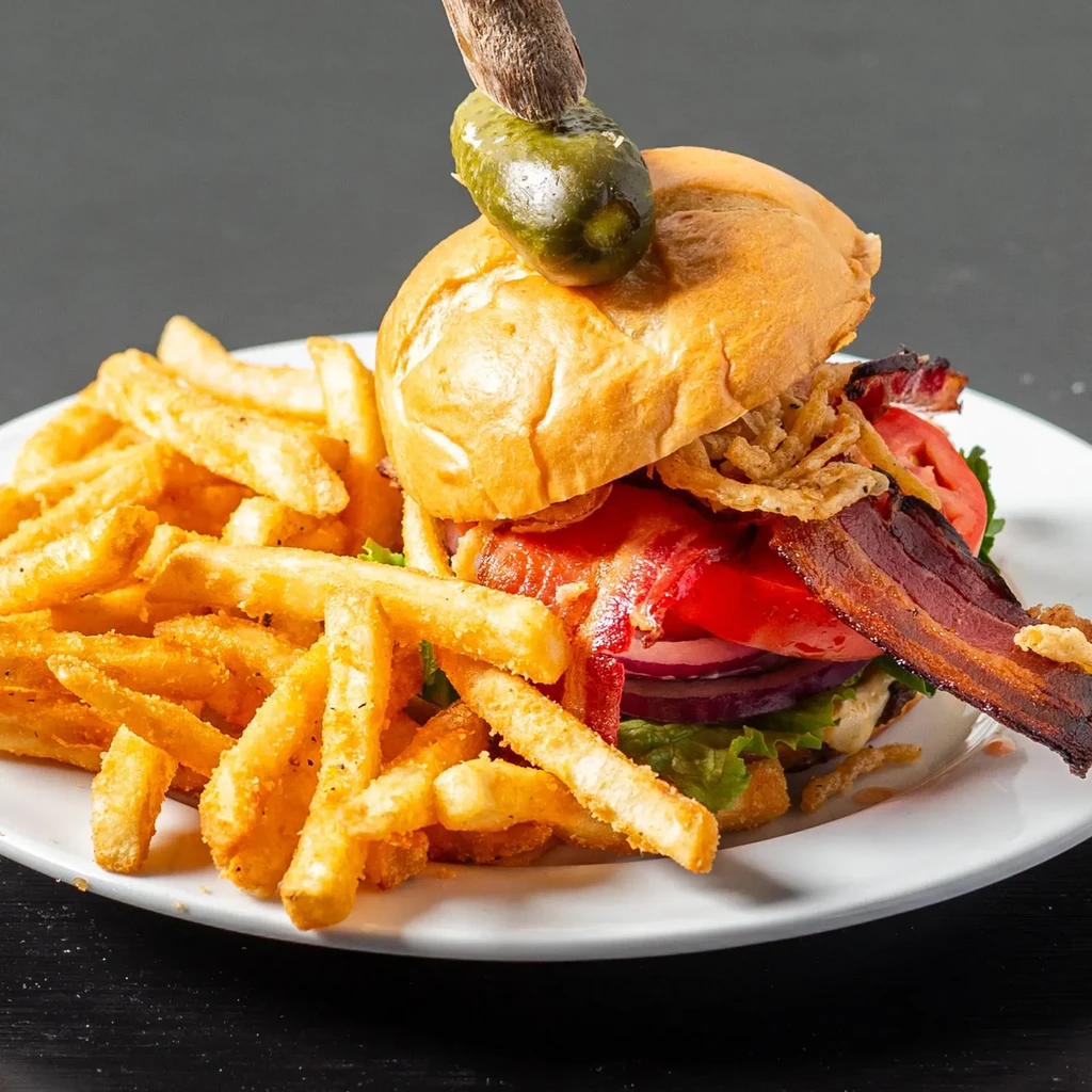 Stacked gourmet burger with bacon, onion rings, pickle, and golden fries.