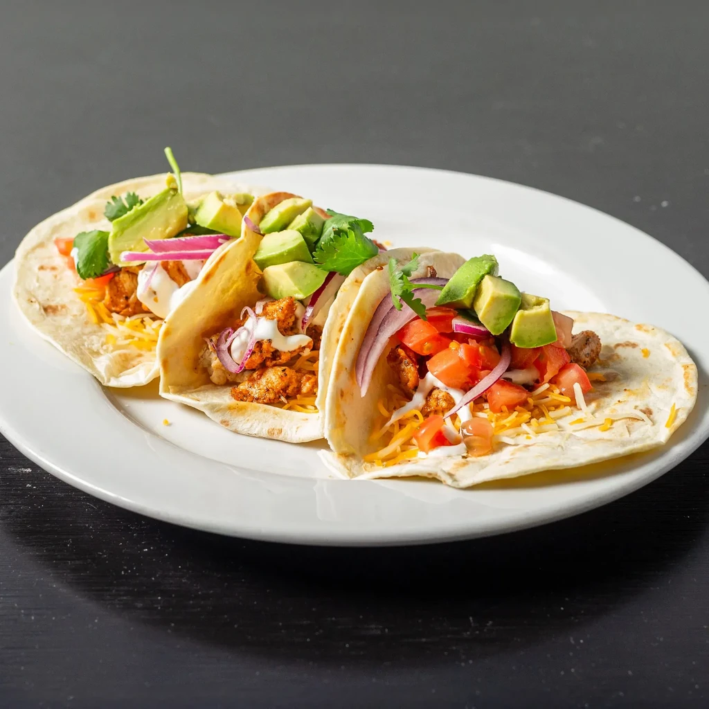 Three tacos with seasoned meat, avocado, cheese, tomatoes, and cilantro.