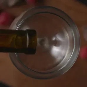 Dark bottle pours liquid into clear wine glass on wood.