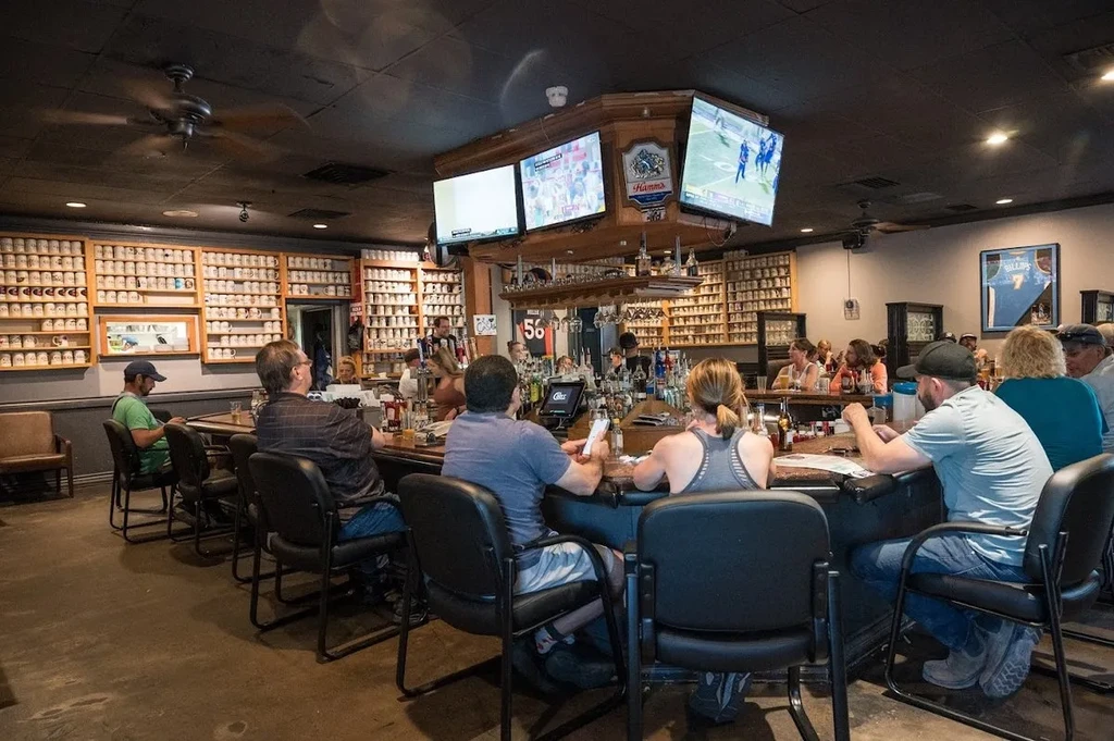 Lively sports bar with patrons, bartenders, and extensive mug display.