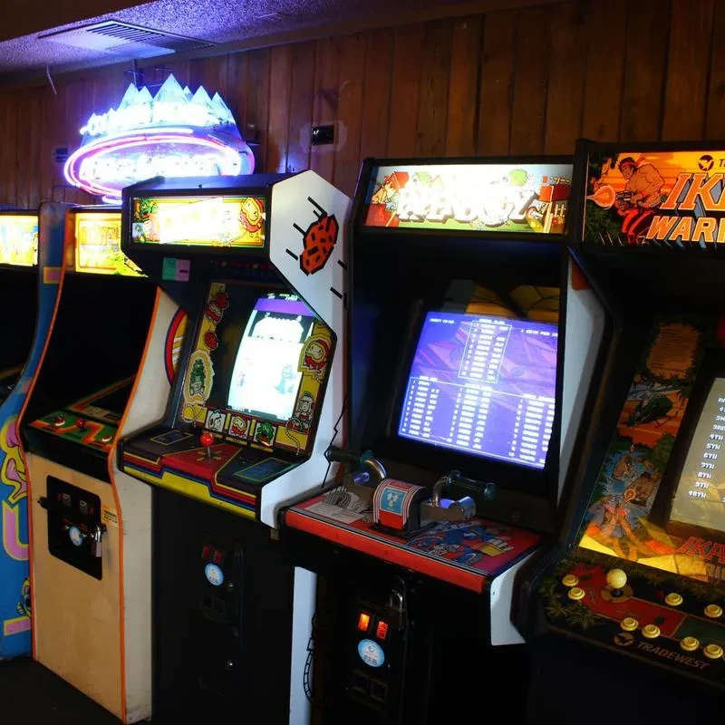 Colorful arcade machines and neon sign glow in a dim barcade.