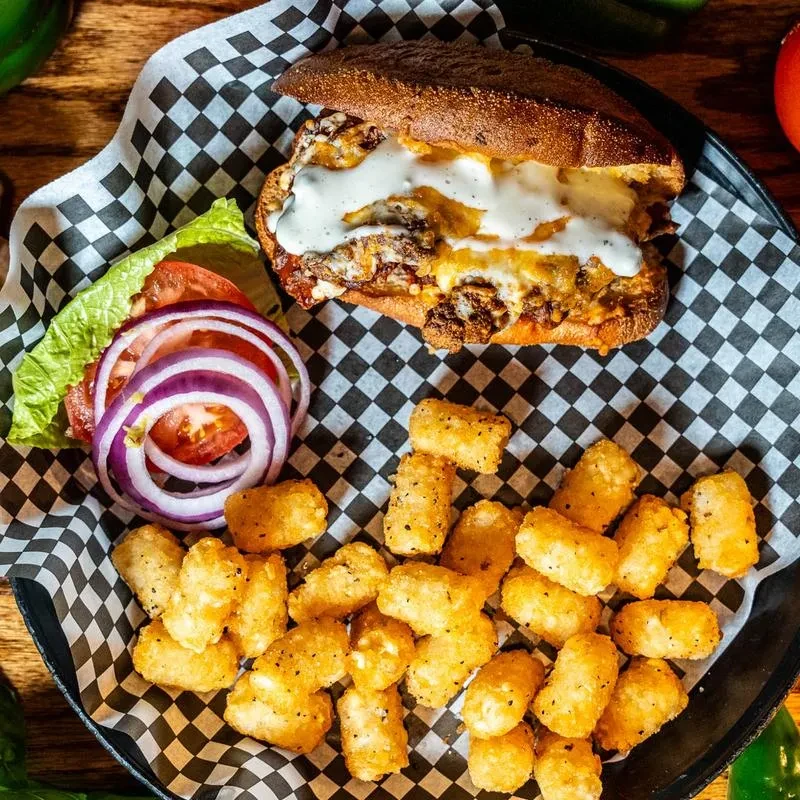 Cheesesteak-style sandwich, tater tots, and fresh vegetables in a black pan.