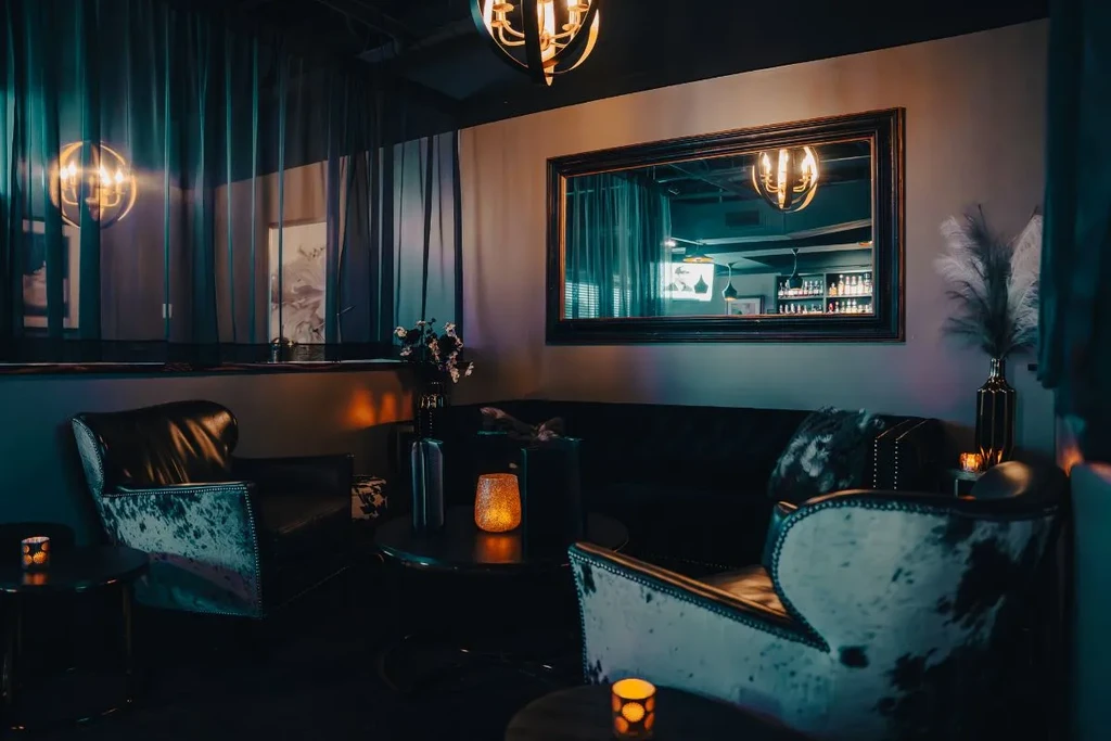 Dimly lit lounge with armchairs, sofa, and mirror reflecting a bar.