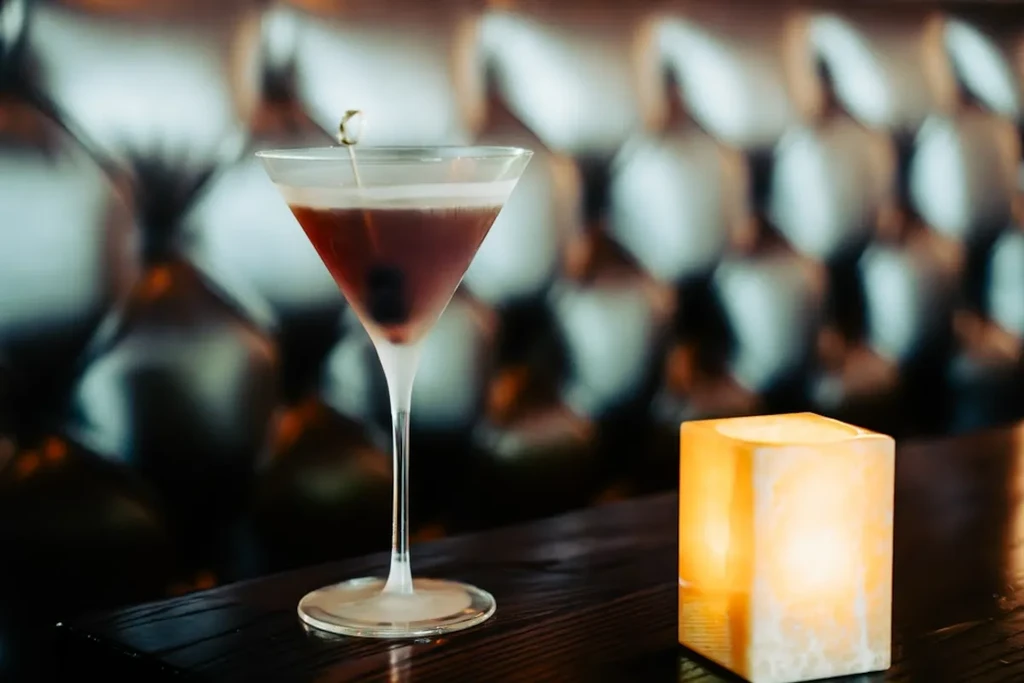Martini glass with a reddish-brown cocktail, cherry, and glowing lamp.