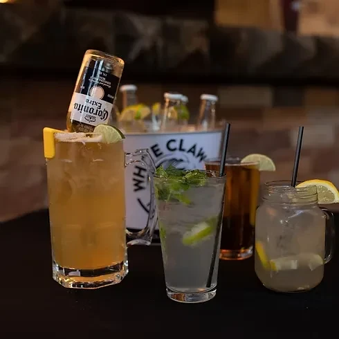 Four garnished alcoholic drinks and a White Claw cooler on a dark surface.