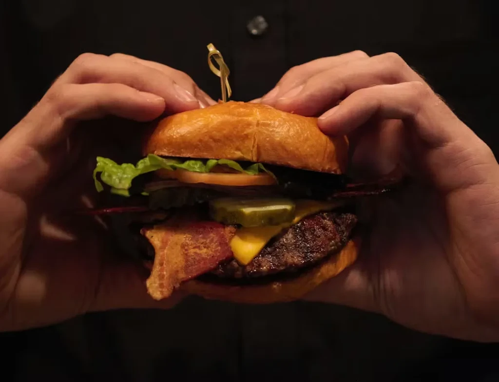 Close-up of a gourmet burger with bacon, cheese, vegetables, held by hands.