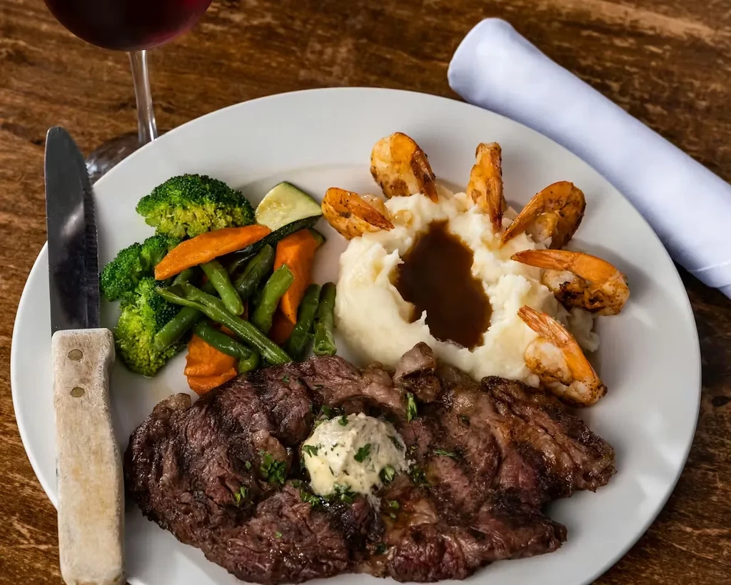 Plated surf and turf dinner: steak, shrimp, vegetables, and mashed potatoes.