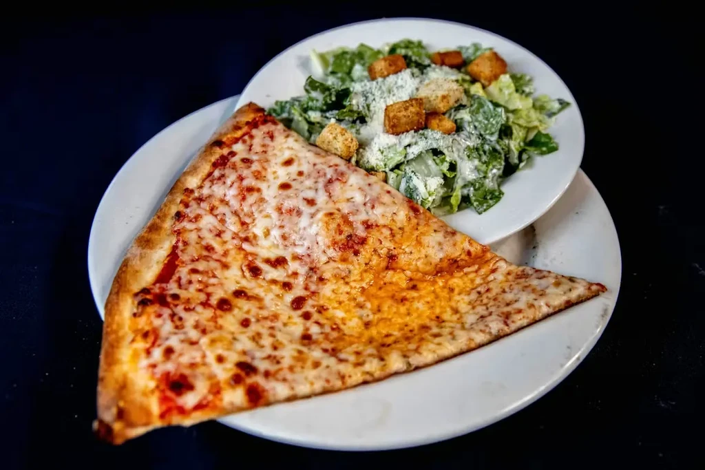 Close-up of cheese pizza slice and Caesar salad on white plates.