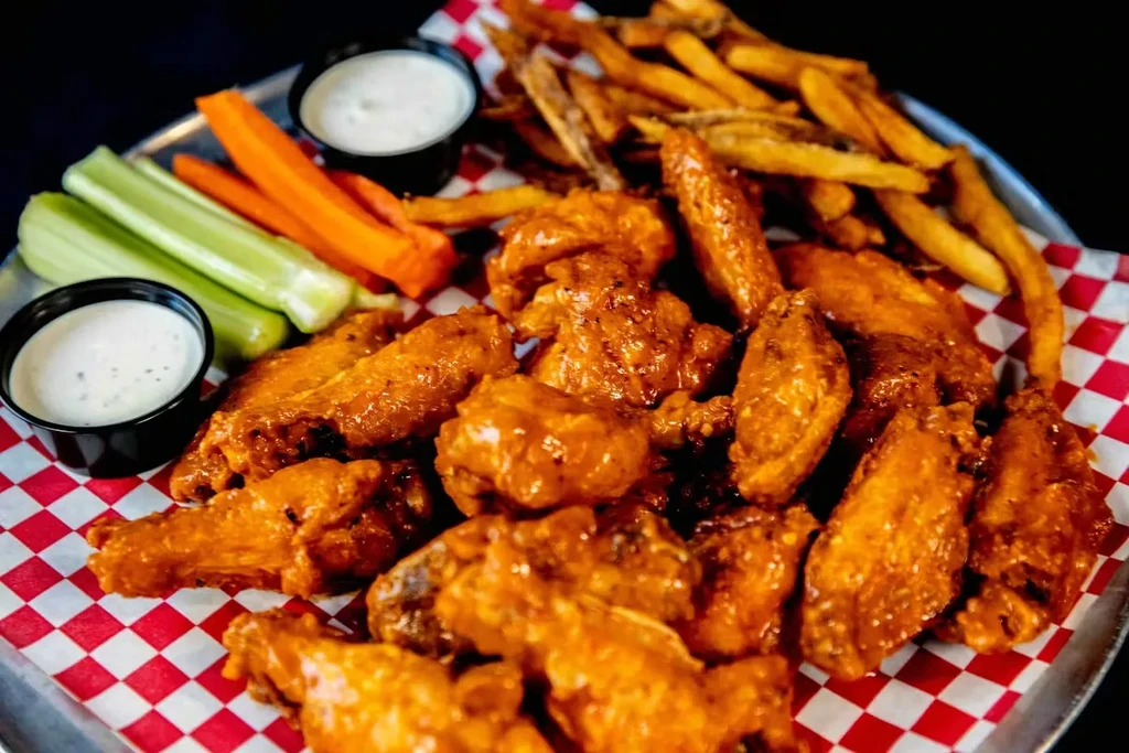 Saucy chicken wings, fries, celery, carrots, and dipping sauce on a platter.