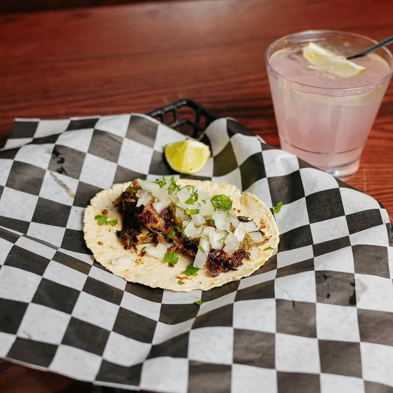 Street taco with meat, onions, cilantro, and pink drink on wood.